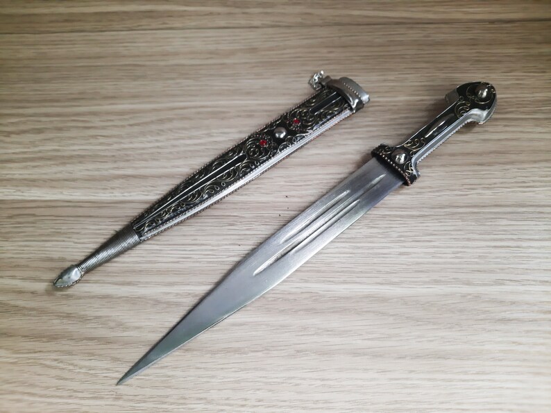 May include: A silver dagger with an ornate handle and a black sheath. The dagger has a sharp point and a double-edged blade.