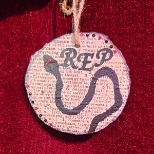 May include: A round, cream-coloured ornament with a black snake wrapped around the word "REP" in black lettering. The ornament is decorated with black and red beads.