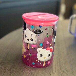 May include: A clear plastic tumbler with a pink lid and base. The tumbler features cartoon character designs, including Hello Kitty, on a pink background with stars and hearts. The tumbler is designed for cold beverages.