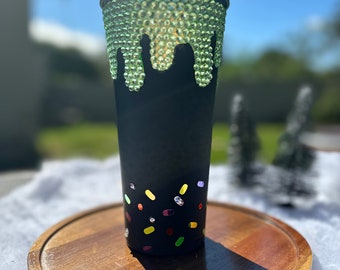 May include: Black tumbler with a green rhinestone drip design and colorful confetti-like accents. The top of the tumbler is also black. The tumbler sits on a wooden tray.