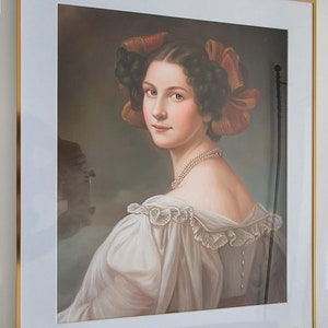 Op de afbeelding: Een ingelijst portret van een vrouw met donker haar en een witte jurk. Ze draagt een rood-oranje lint in haar haar en een parelketting. Het portret is tegen een bruine achtergrond.
