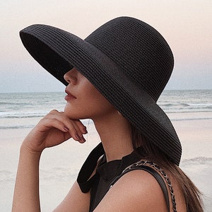 May include: A black sun hat with a wide brim. The hat is made of woven material and has a ribbon detail. The hat is worn on the beach. The hat is a stylish accessory for sun protection.