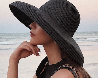 Puede incluir: Un sombrero negro para el sol con ala ancha. El sombrero está hecho de material tejido y tiene un detalle de cinta. El sombrero se usa en la playa. El sombrero es un accesorio elegante para la protección solar.
