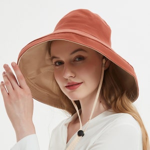 May include: A terracotta-colored sun hat with a wide brim and a beige underside. The hat features a chin strap with a black toggle. The hat is designed to provide sun protection and is suitable for outdoor activities.