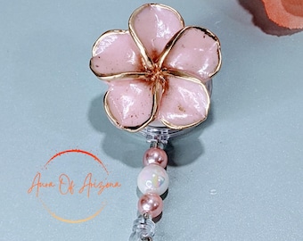 Puede incluir: Carrete de identificación en forma de flor rosa con detalles dorados y un cordón retráctil de plástico transparente. El carrete está decorado con cuentas rosas y blancas.