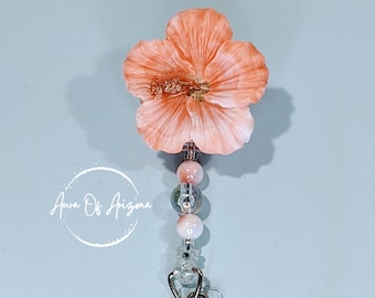 May include: A badge reel featuring a peach-colored hibiscus flower topper with a textured center. Below the flower, a vertical strand of beads in shades of peach and translucent gray connects to a clear plastic strap with a metal snap button.