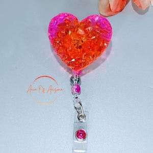 May include: A heart-shaped badge reel with a gradient of pink and orange glitter. The reel has a clear plastic clip with a pink gem and a beaded accent. The text "Anna Of Arizona" is in the image.
