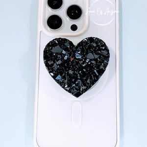 May include: A white smartphone case featuring a black heart-shaped phone grip with a textured, crystalline surface. The grip is centered on the back of the case, which has a camera cutout at the top. The text Aura Of Arizona is visible.
