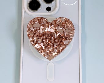 May include: A white smartphone case featuring a heart-shaped phone grip with a rose gold, textured, faux-crystal surface. The phone case has a triple-lens camera cutout and the text Aura Of Arizona printed in a white circular logo.