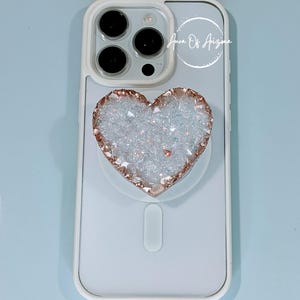 May include: A white smartphone case with a triple-lens camera and a heart-shaped phone grip featuring a textured, iridescent white druzy surface with a metallic rose gold edge. The text Aura Of Arizona is printed in white near the top right.