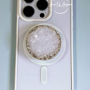 May include: A light purple smartphone case with a white border and a circular, textured, silver-toned glittery grip attachment centered on the back. The text Aura Of Arizona is printed in white script within a circular outline near the camera lens.