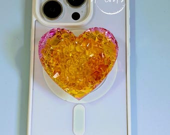 May include: A white smartphone case with a triple-lens camera cutout and a heart-shaped phone grip featuring a faceted, amber-yellow and pink crystalline texture. The text Aura Of Arizona is printed in white script within a circular outline.