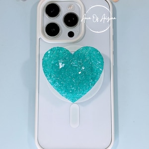 May include: A white smartphone case featuring a textured, heart-shaped teal attachment with a crystalline surface. The case has a triple-lens camera cutout and the text Aura Of Arizona printed in white near the top right corner.