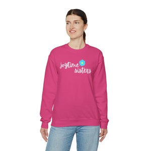 May include: A magenta crew neck sweatshirt featuring the words "joytime sisters" in white script, with a blue and yellow flower design. The garment is a vibrant magenta colour.