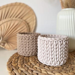 May include: Two crocheted baskets, one beige and one light brown, are sitting on a woven straw mat. The baskets are made of thick rope and have a textured, woven pattern.