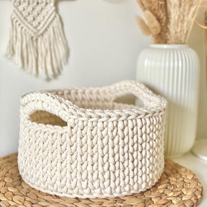 May include: A cream-coloured crocheted basket with two handles sits on a woven straw mat. The basket is made of thick, textured yarn and has a rounded shape.