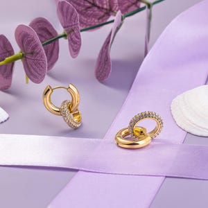 May include: A pair of gold hoop earrings with a smaller hoop attached to the larger hoop. Both hoops are encrusted with small clear stones.