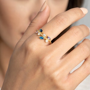 May include: A gold-colored ring in a leaf design, adorned with colorful gemstones. The ring features a branch-like structure with various colored stones, including blue, purple, and white, set along its length. The ring is worn on a finger.