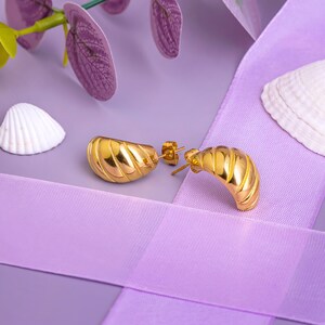 May include: A pair of gold hoop earrings with a textured, wavy design. The earrings are displayed on a purple background with a white seashell.