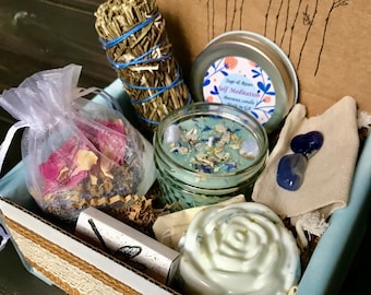 May include: A gift box containing a sage bundle tied with blue string, a tin candle labeled Sage et Roses Self Meditation, a glass jar candle with botanical toppings, a white rose-shaped soap, two blue stones, and a sheer bag of dried flowers.