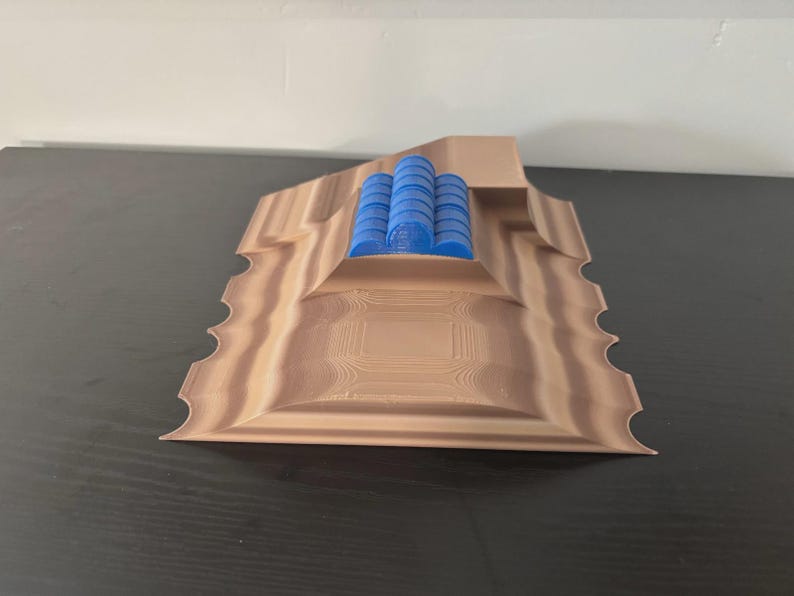May include: A 3D-printed architectural model in shades of brown and blue. The model features a layered design with a central raised section holding several blue cylindrical structures. The edges have a decorative, scalloped pattern.
