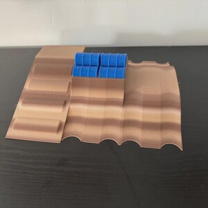 May include: A 3D-printed model of a desert landscape with a series of brown, layered terrain features. Four small blue barrels are positioned on the highest point of the landscape. The model is designed for tabletop gaming or display.