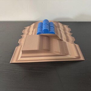 May include: A 3D-printed model of a landscape with a stepped structure and wavy edges. The model is primarily tan with blue cylindrical structures on top. The design appears to be a miniature architectural or terrain representation.