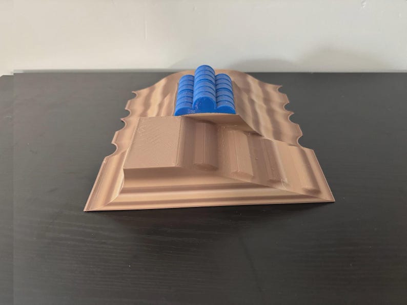 May include: A 3D-printed model of a landscape with a stepped structure and wavy edges. The model is primarily tan with blue cylindrical structures on top. The design appears to be a miniature architectural or terrain representation.