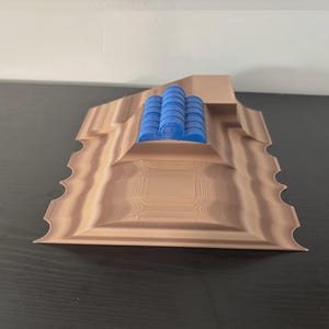 May include: A 3D-printed sculpture featuring a layered, light brown base with a textured surface and scalloped edges. Three blue cylindrical objects are stacked on top. The sculpture is displayed on a dark surface.