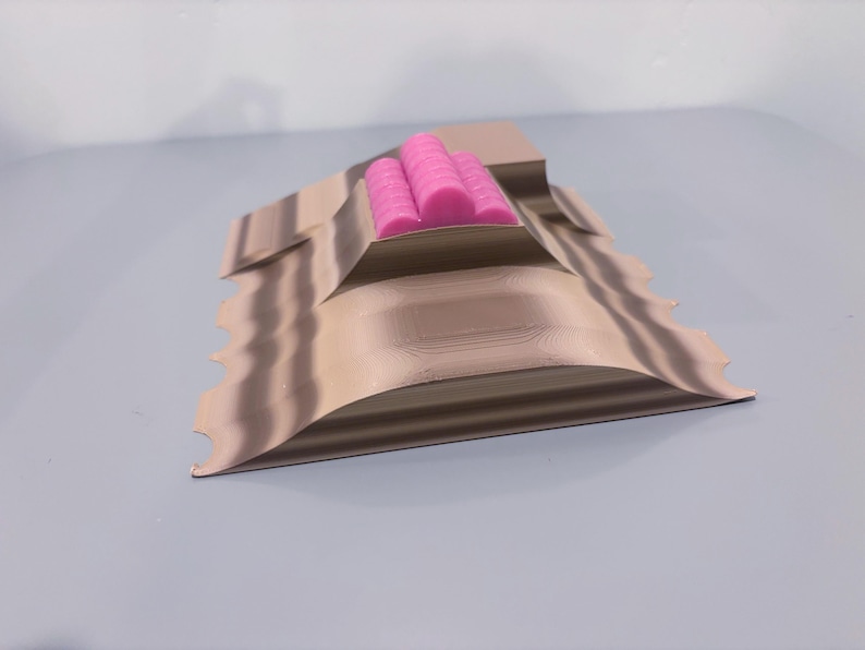 May include: A tan, layered, 3D-printed sculpture with a wavy edge. Three bright pink cylindrical objects are nestled in the center. The sculpture is on a light gray surface.