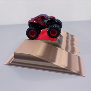 May include: A red and black monster truck toy with large black tires and red rims is positioned on a tan ramp. The ramp has a red platform at the top and a wavy design on the side.