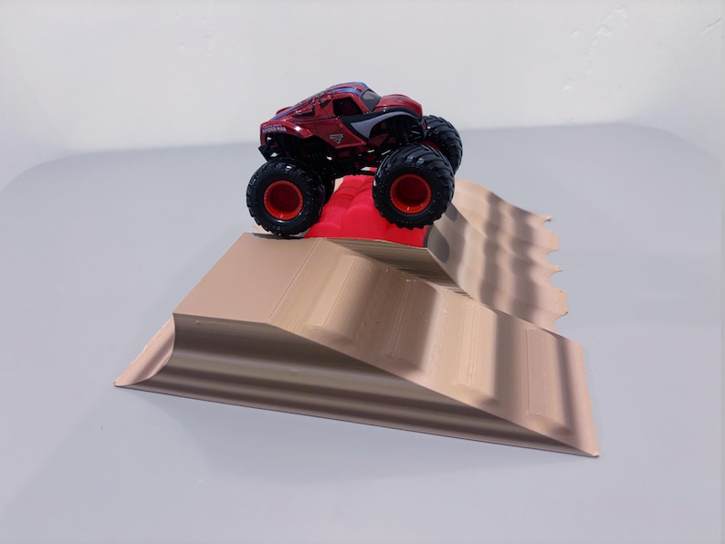 May include: A red and black monster truck toy with large black tires and red rims is positioned on a tan ramp. The ramp has a red platform at the top and a wavy design on the side.