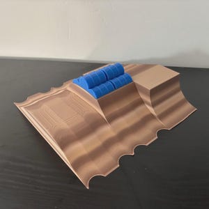 May include: A 3D-printed model featuring a brown, layered landscape with a series of blue cylindrical objects. The model has a textured surface and a scalloped edge. The design is geometric and modern.