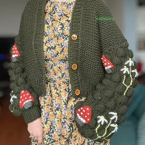 May include: A green knitted cardigan with mushroom and daisy appliques. The cardigan has a button closure and long sleeves. The text "Yesimdesign" is visible on the cardigan.