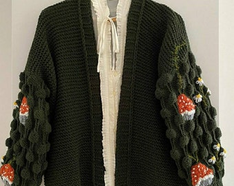 Pode incluir: Um casaco de malha verde escuro com uma camisa branca de botões por baixo. O casaco tem uma malha texturizada e apresenta cogumelos e margaridas bordadas nas mangas.