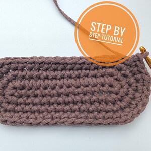 K&ouml;nnte beinhalten: Nahaufnahme einer braunen geh&auml;kelten ovalen Form mit einer goldenen H&auml;kelnadel und einem Garnfaden. Der Text "STEP BY STEP TUTORIAL" ist in einem Kreis sichtbar.