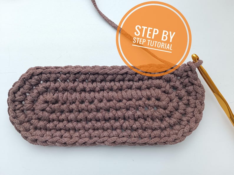 K&ouml;nnte beinhalten: Nahaufnahme einer braunen geh&auml;kelten ovalen Form mit einer goldenen H&auml;kelnadel und einem Garnfaden. Der Text "STEP BY STEP TUTORIAL" ist in einem Kreis sichtbar.