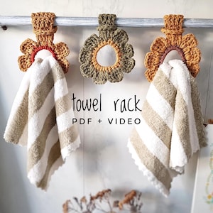 May include: Three crocheted flower towel racks in shades of brown, green, and orange hang on a white towel bar. The towel racks are shaped like flowers with a ring in the center to hold a towel. The text "towel rack PDF + VIDEO" is printed below the towel racks.