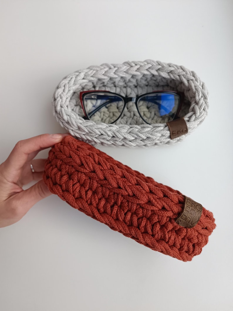 May include: Two crocheted baskets, one gray and one rust colored. The gray basket has a pair of glasses inside. Both baskets have a leather tag with the word "EKO" on them.