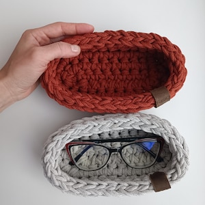 May include: Two crocheted oval-shaped baskets, one in a rust orange color and the other in a light gray color. The gray basket has a pair of eyeglasses inside.