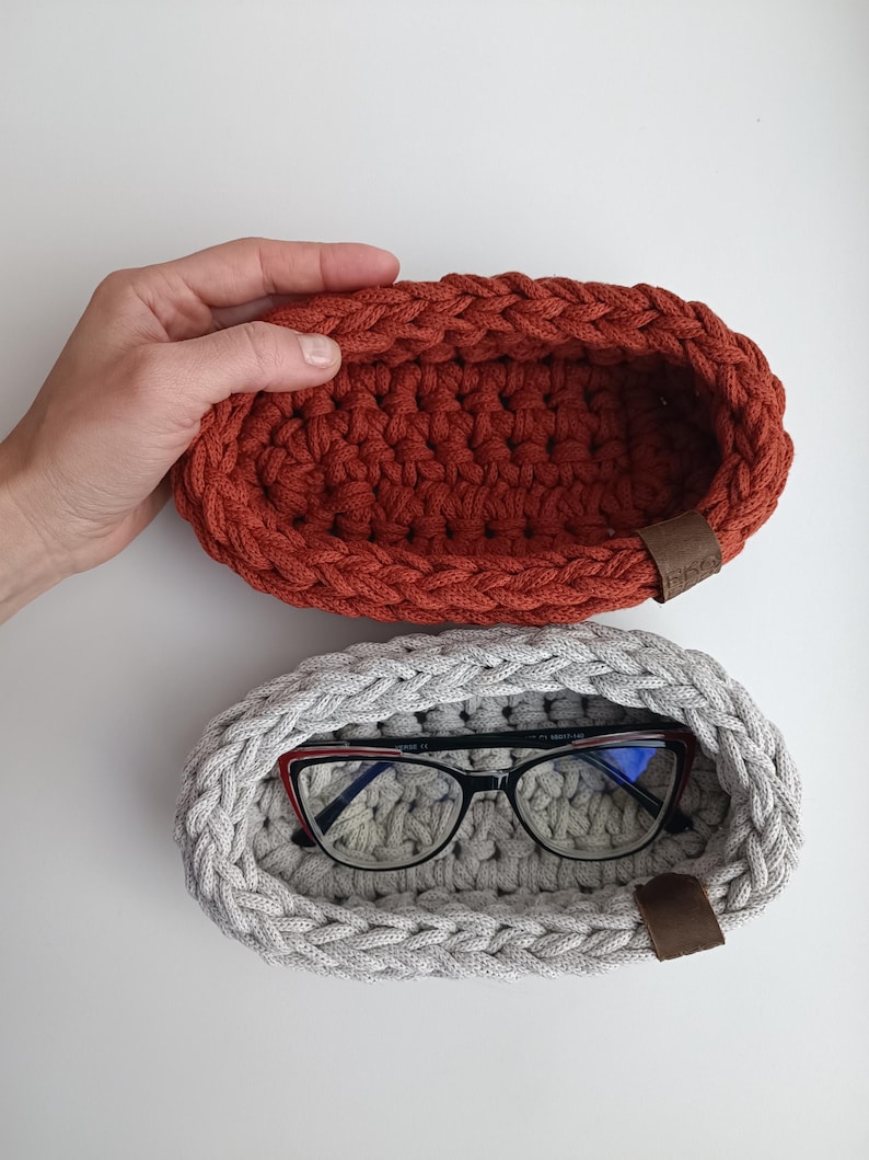 May include: Two crocheted oval-shaped baskets, one in a rust orange color and the other in a light gray color. The gray basket has a pair of eyeglasses inside.