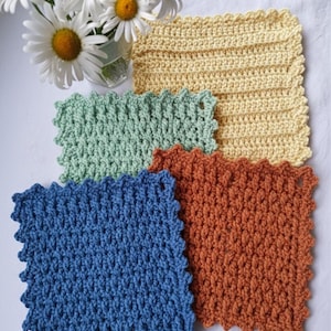 Könnte beinhalten: Vier handgefertigte gehäkelte Waschlappen in Blau, Grün, Gelb und Orange. Jeder quadratische Lappen hat ein strukturiertes Muster und einen gewellten Rand. Im Hintergrund steht eine kleine Vase mit Gänseblümchen.