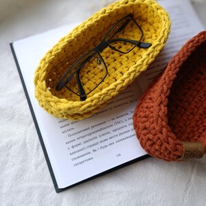 May include: Two crocheted bowls, one yellow and one brown, with a pair of eyeglasses resting in the yellow bowl. The bowls are on top of a book.