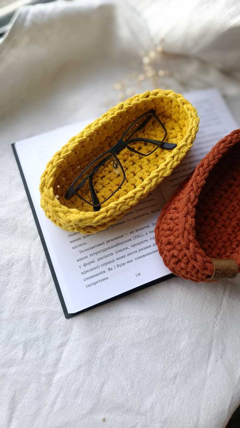 May include: Two crocheted bowls, one yellow and one brown, with a pair of eyeglasses resting in the yellow bowl. The bowls are on top of a book.