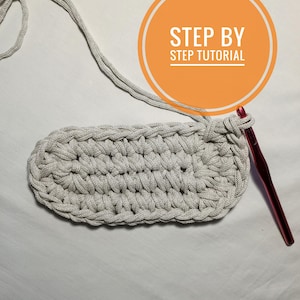 May include: A close-up of a crocheted oval rug in a light gray color. A pink crochet hook is lying on a white surface next to the rug. The text "STEP BY STEP TUTORIAL" is written in a circle on the image.