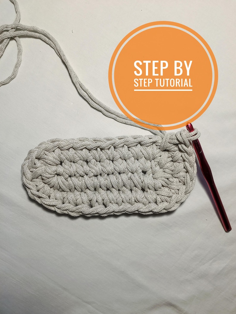 May include: A close-up of a crocheted oval rug in a light gray color. A pink crochet hook is lying on a white surface next to the rug. The text "STEP BY STEP TUTORIAL" is written in a circle on the image.