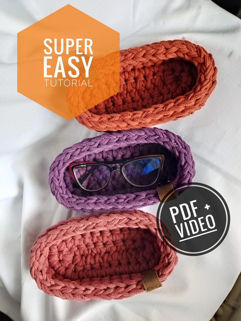 May include: Three crocheted oval-shaped baskets in different colors: orange, purple, and pink. The baskets are made with thick yarn and have a simple design. The text "SUPER EASY TUTORIAL" and "PDF + VIDEO" are visible in the image.
