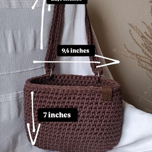 K&ouml;nnte beinhalten: Eine braune geh&auml;kelte Tragetasche mit einer H&ouml;he von 17,8 cm, einer Breite von 23,9 cm und einer Riemenl&auml;nge von 56,9 cm. Die Tasche hat ein braunes Leder-Etikett mit dem Text "SHOP LOCAL" darauf.