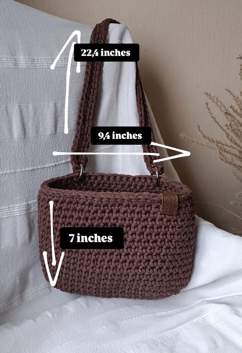 K&ouml;nnte beinhalten: Eine braune geh&auml;kelte Tragetasche mit einer H&ouml;he von 17,8 cm, einer Breite von 23,9 cm und einer Riemenl&auml;nge von 56,9 cm. Die Tasche hat ein braunes Leder-Etikett mit dem Text "SHOP LOCAL" darauf.