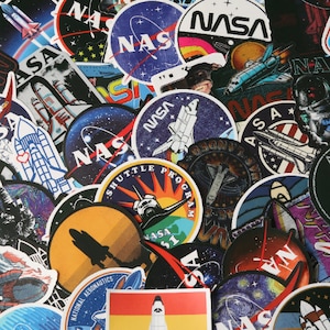 May include: A selection of colourful NASA-themed stickers. The stickers feature various designs, including the NASA logo, space shuttles, astronauts, and celestial imagery. The stickers are round and rectangular, with a variety of colours and styles.